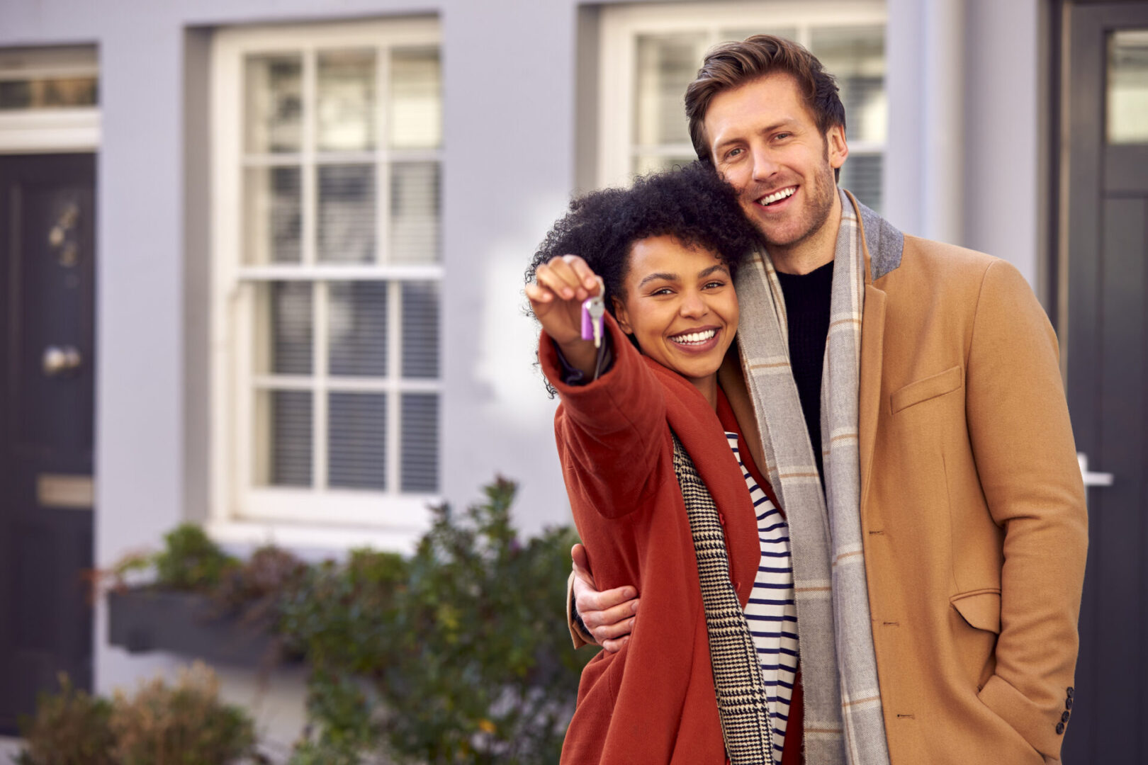 Portrait Of Multi Cultural Couple Outdoors On Moving Day Holding Keys To New Home In Fall Or Winter Couple smiling, holding house keys outside home.